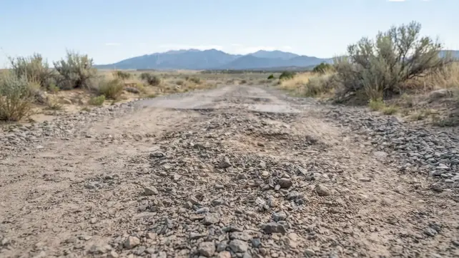 Close-up of a rural gravel driveway showing ruts, tire impressions, uneven aggregate, and surface deterioration in Costilla County Colorado