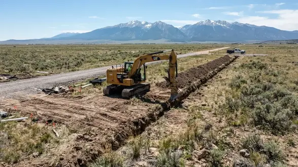 Tracked excavator installing a utility trench across varied rural ground in Costilla County